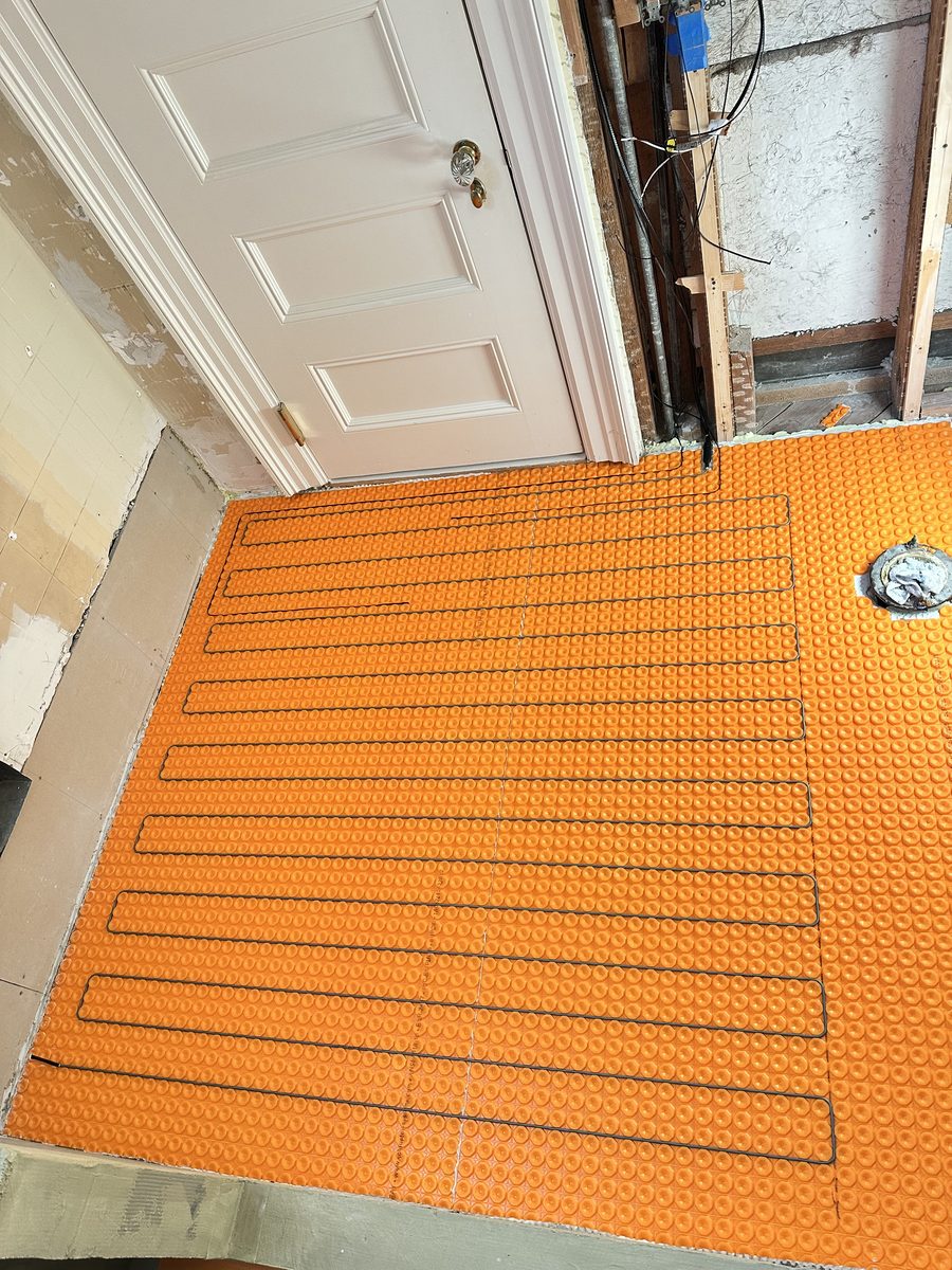 Orange uncoupling membrane laid over a bathroom subfloor with electric radiant heating cable run through it, before any tile goes down