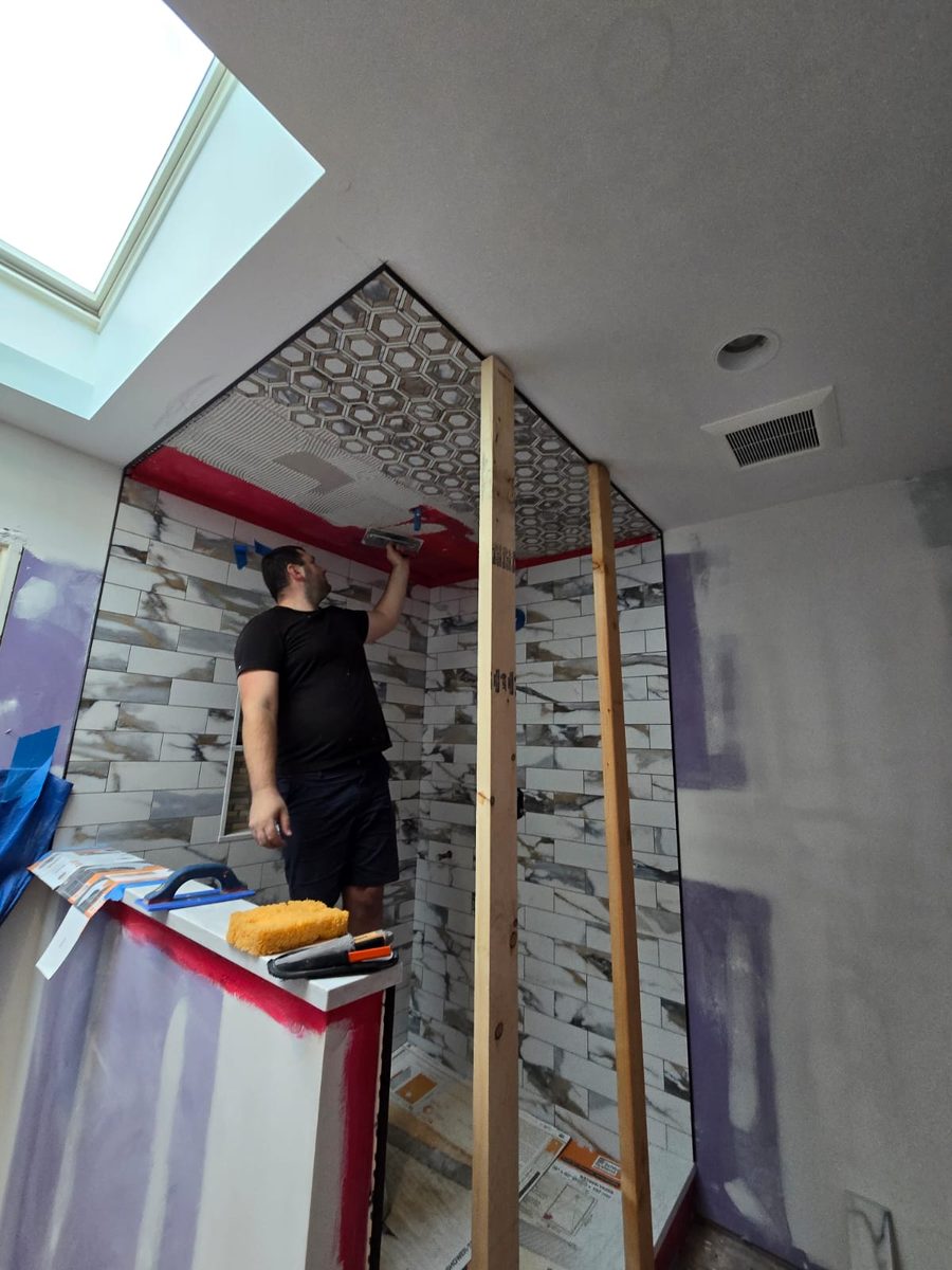 Magic of Tiles installer combing thinset onto a shower ceiling, with marble look subway tile on the walls and red waterproofing membrane visible at the edges